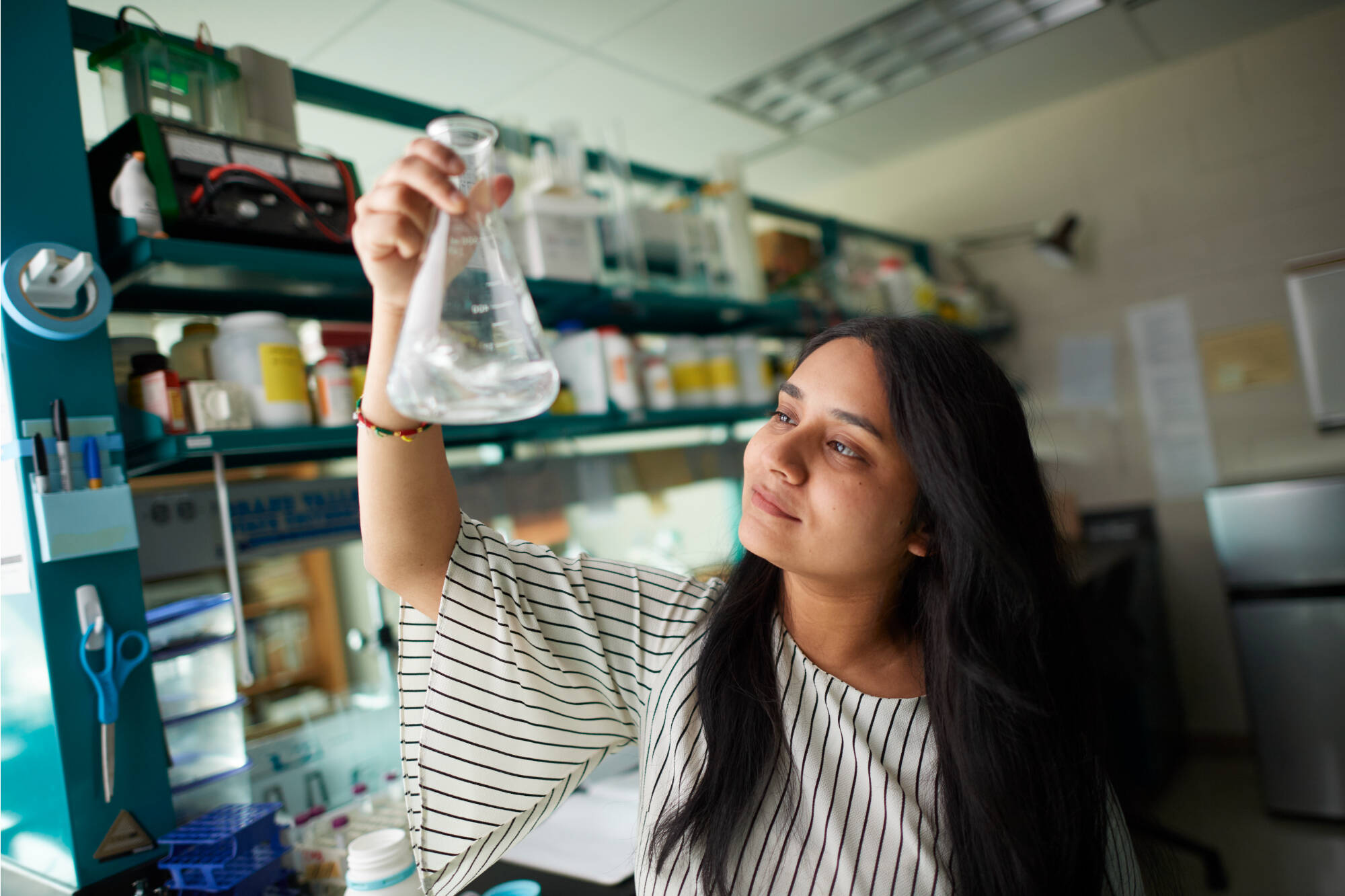 Honors student, Devina Chauhan, working in a lab, looking up at a solution inside a beaker she is holding.